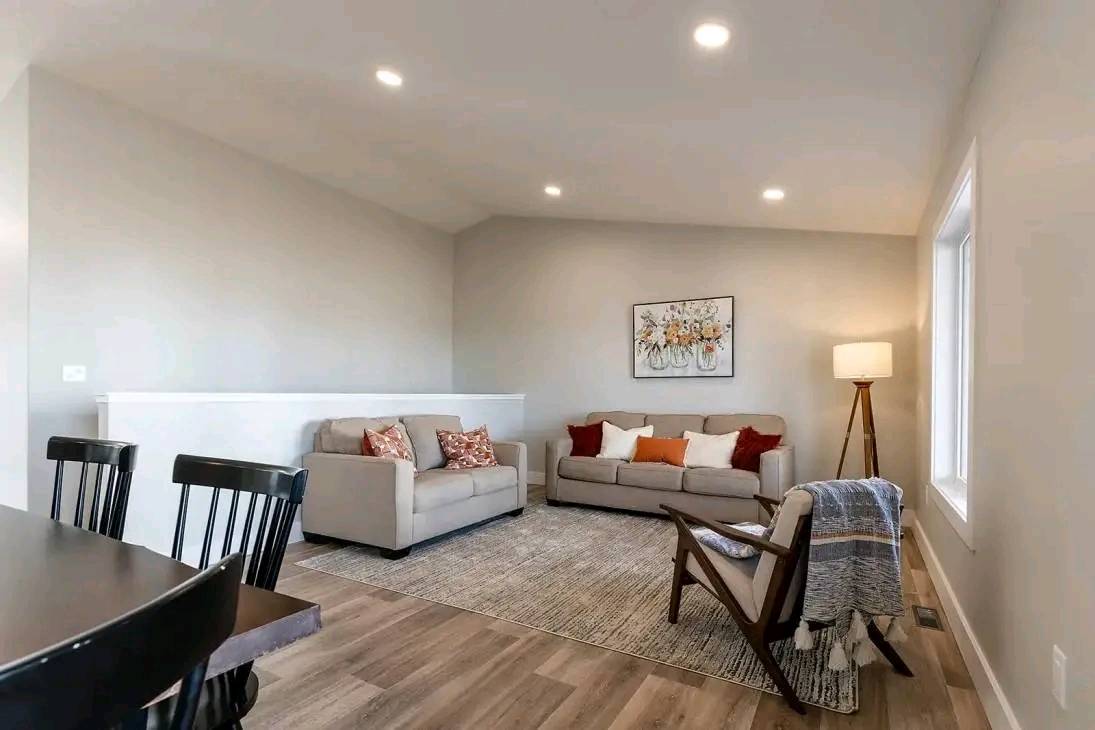 A cozy living room with neutral tones features two beige sofas, colorful cushions, a standing lamp, and wall art. A wooden chair with a blanket and a dining table with chairs add warmth and elegance.