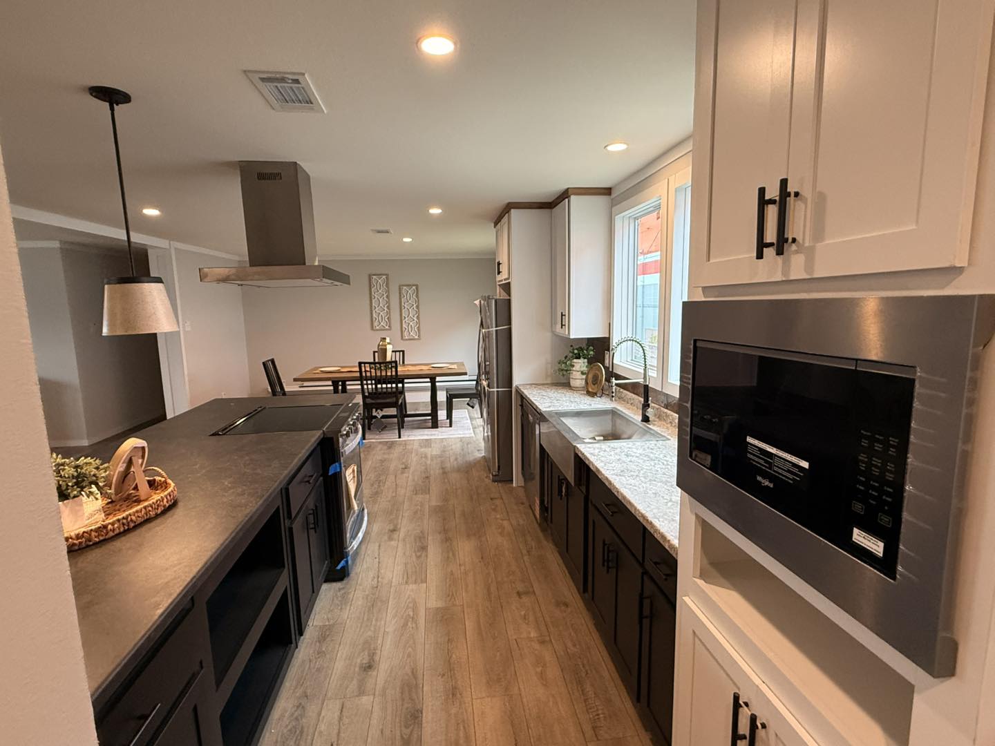 Modern kitchen with a long granite counter, dark cabinets, and stainless steel appliances. A dining table is visible in the bright, open layout.