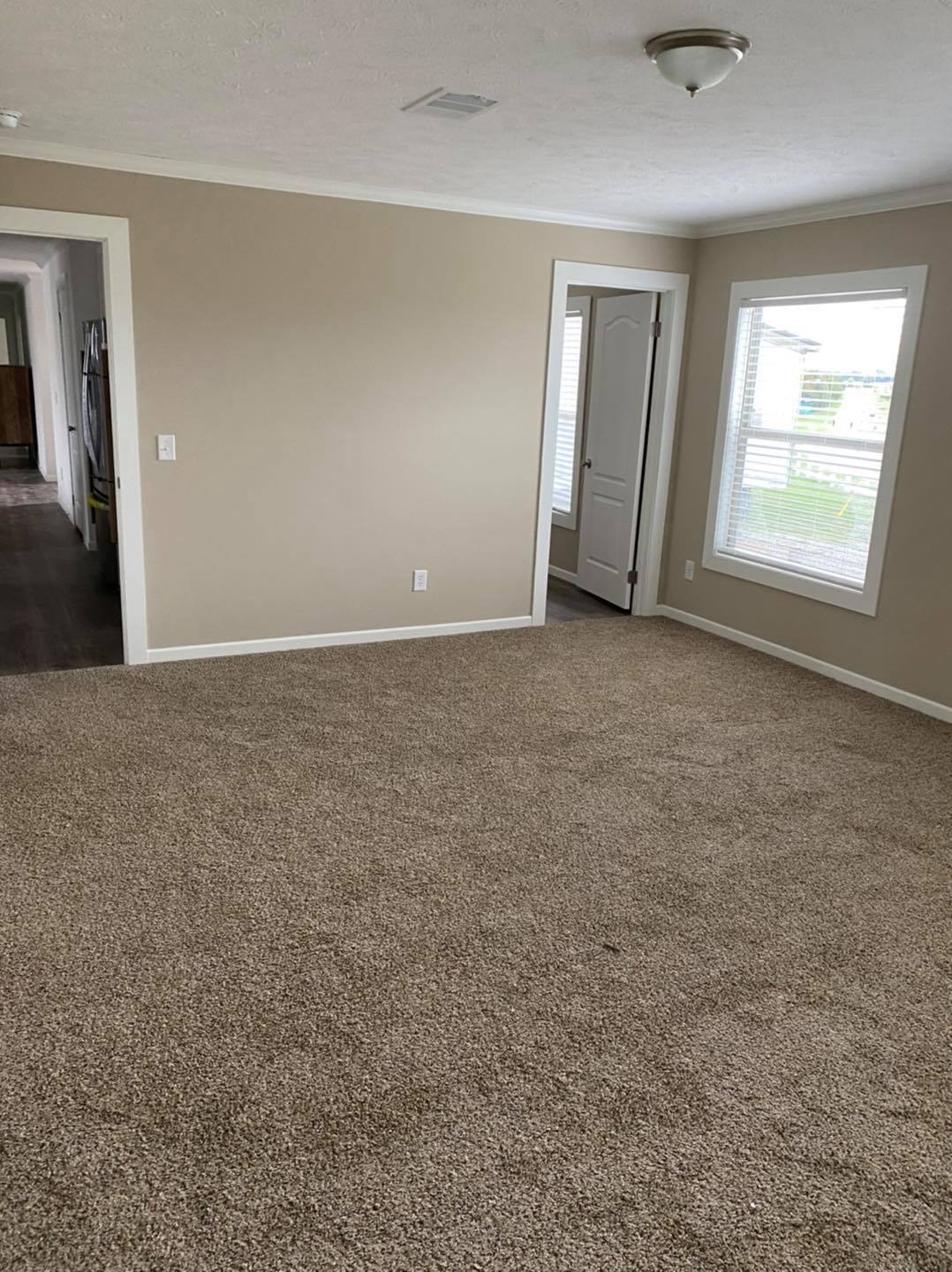Spacious empty room with beige walls and carpet, two large windows on the right, a white door in the corner, and a hallway leading to another room. Natural light adds warmth.