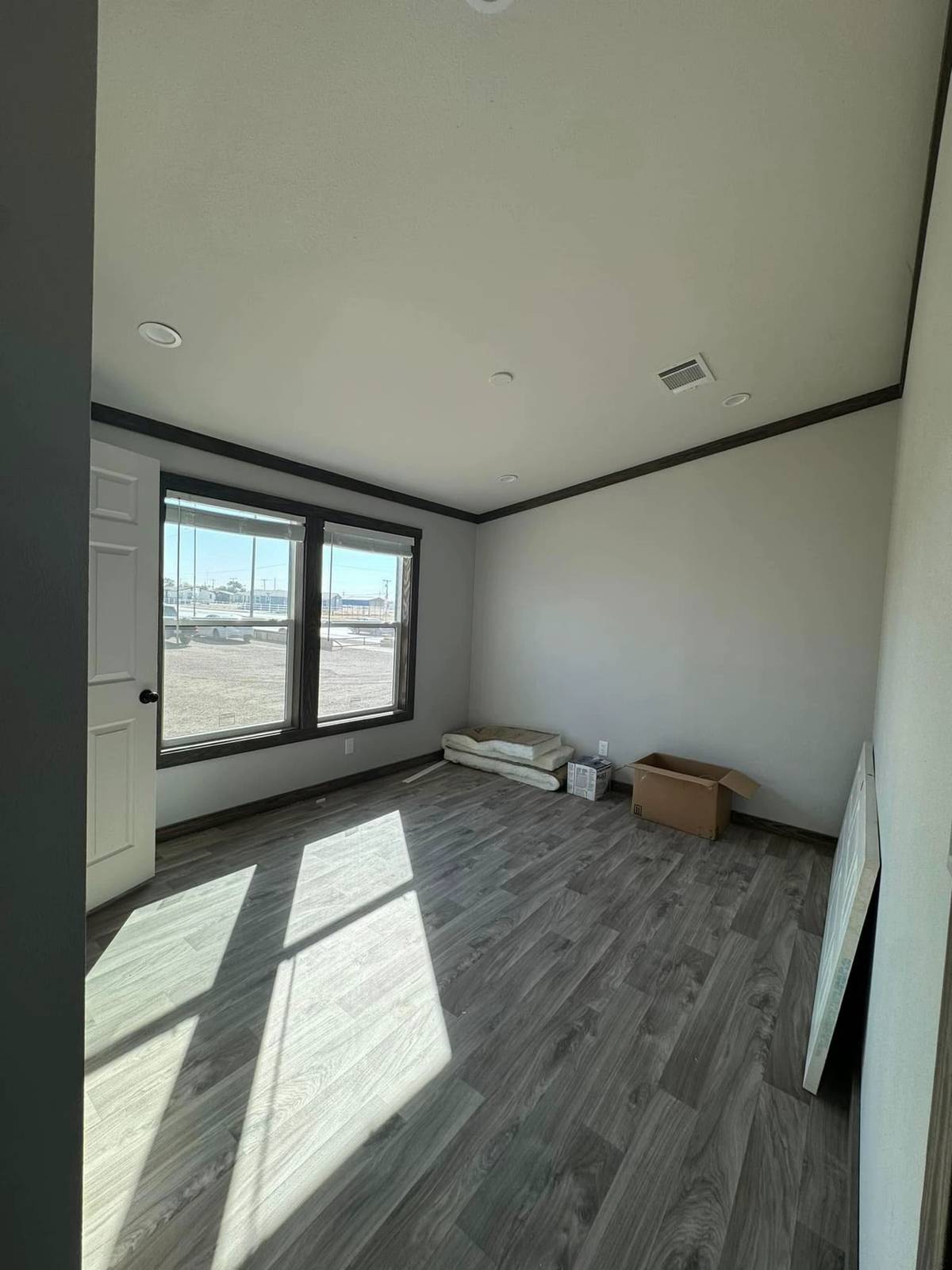 Empty room with light gray walls and wood flooring, lit by sunlight through three large windows. Unpacked boxes and clutter sit in one corner.