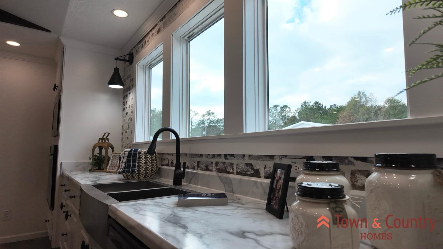 Modern kitchen with a sleek marble countertop, black fixtures, decorative jars, and a view of trees through large windows, creating a bright, airy feel.