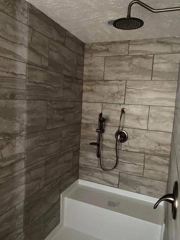 A modern bathroom features a walk-in shower with marble-patterned gray tiles. It has a wall-mounted showerhead and dark bronze fixtures. The tone is sleek and contemporary.