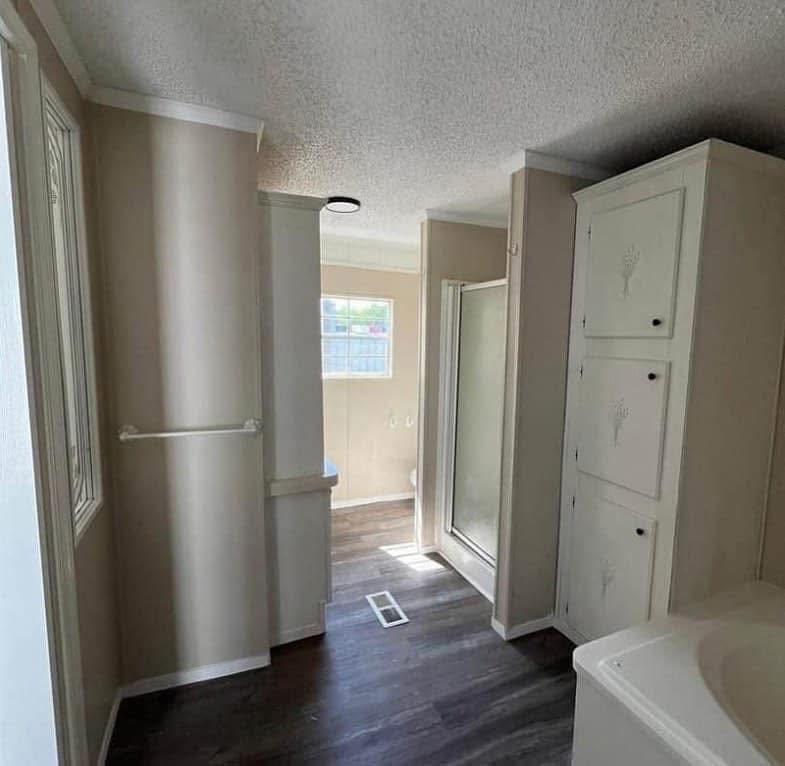 Narrow bathroom with wooden floor, white walls, and cabinets. Shower stall and sink visible. Natural light from window creates a calm atmosphere.