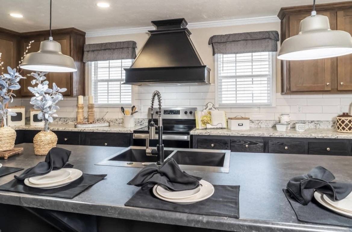 Modern kitchen with a black island, pendant lights, and a center sink. Elegant table settings with black napkins, wooden cabinets, and a stainless steel stove.