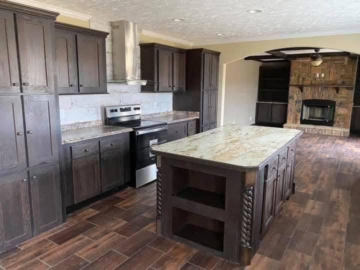 A spacious kitchen features dark wood cabinets, a granite island with ornate pillars, and a stainless steel stove. The adjacent room has a fireplace and wooden floors, creating a warm, elegant atmosphere.