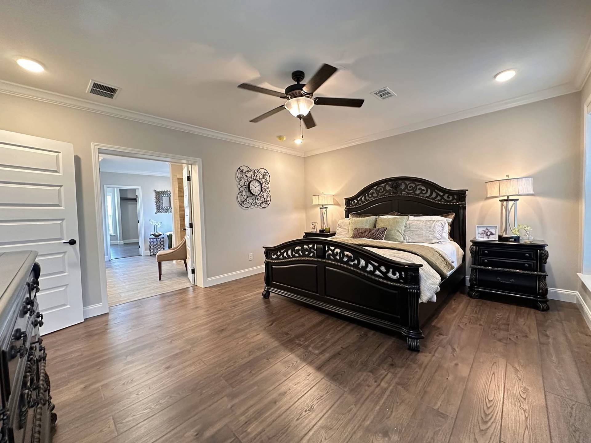 Spacious bedroom with dark wood furniture, including a large ornate bed and nightstands. Warm lighting, ceiling fan, and a wall clock create a cozy ambiance.