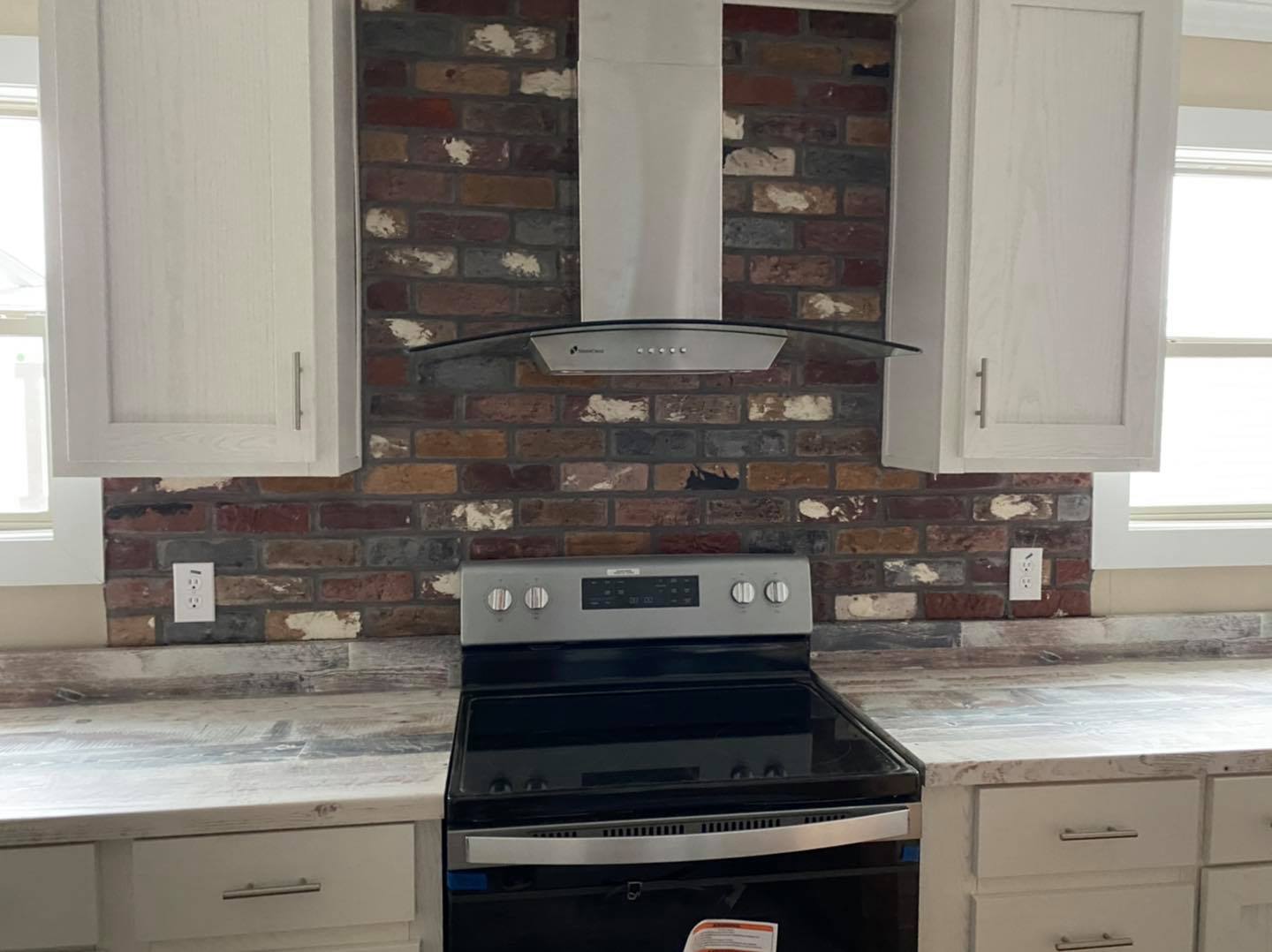 Modern kitchen with a brick backsplash, stainless steel range with black cooktop, and silver hood. White cabinets flank the stove, creating a rustic yet sleek look.
