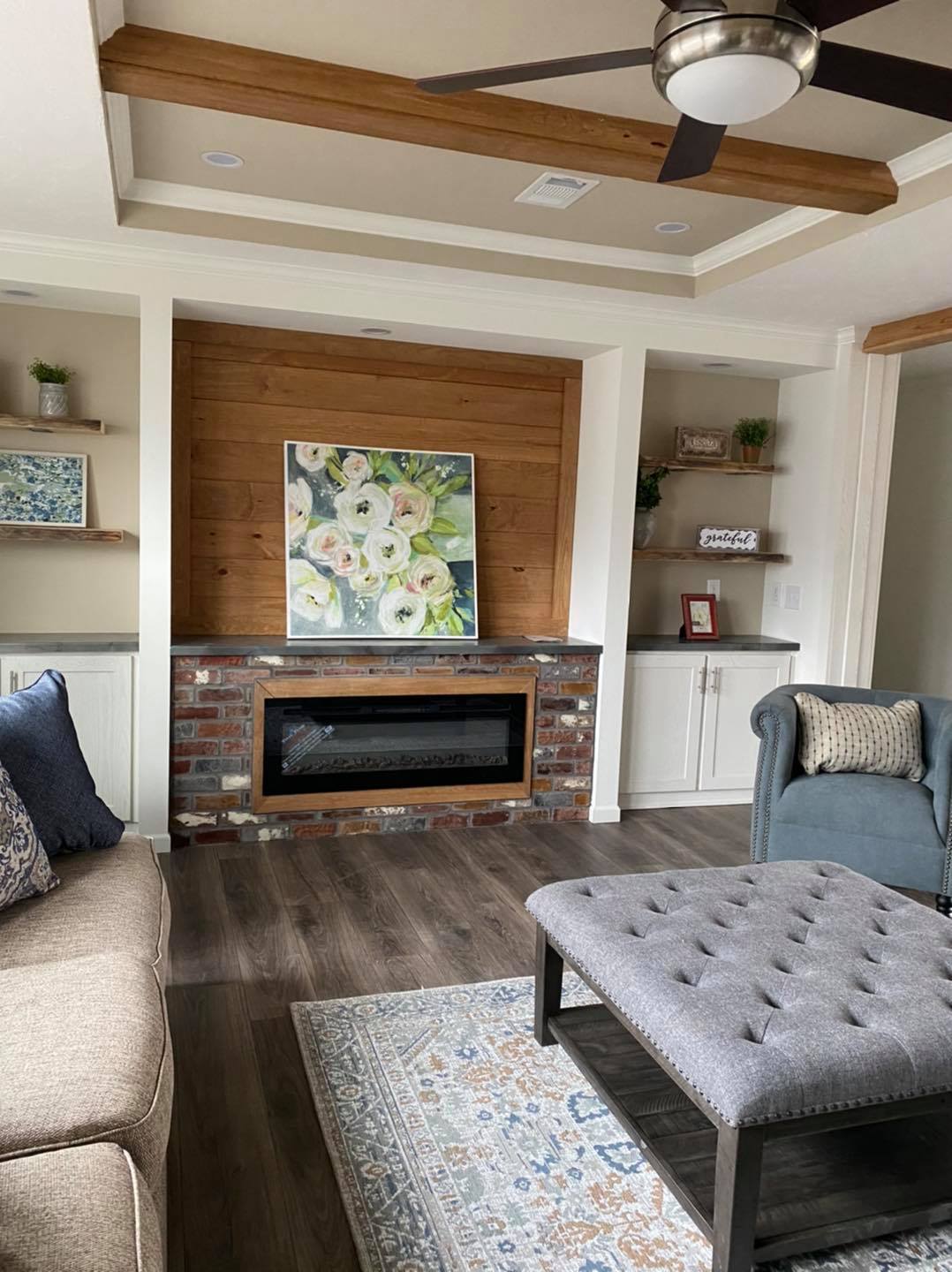 A cozy living room with a modern, rustic fireplace. A floral painting is above the brick mantel. Neutral tones, wood beams, and soft cushions add warmth.