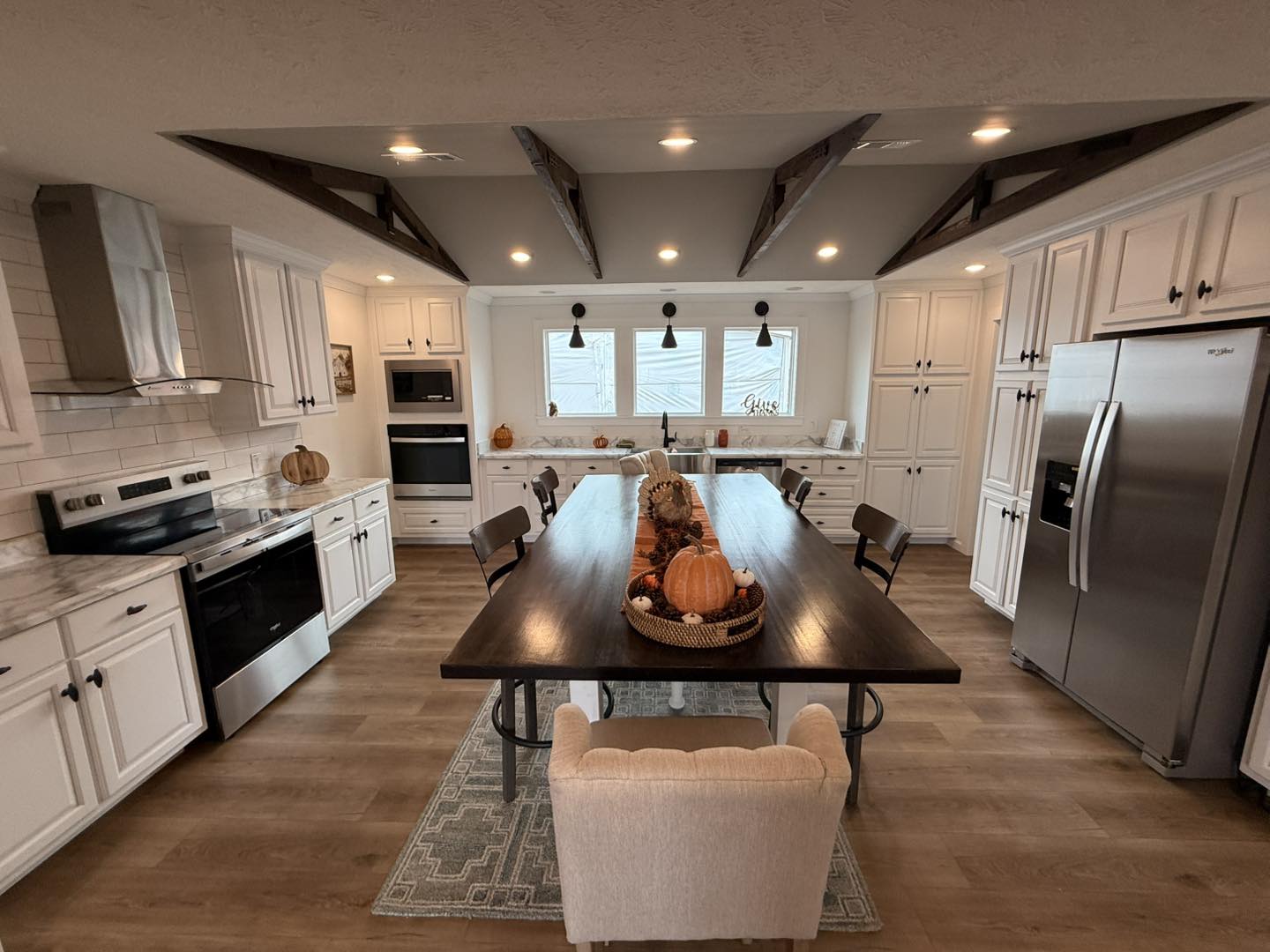Spacious kitchen with white cabinets and marble countertops. Central wooden table with fall decorations and pumpkins. Warm, cozy ambiance with wood floors.