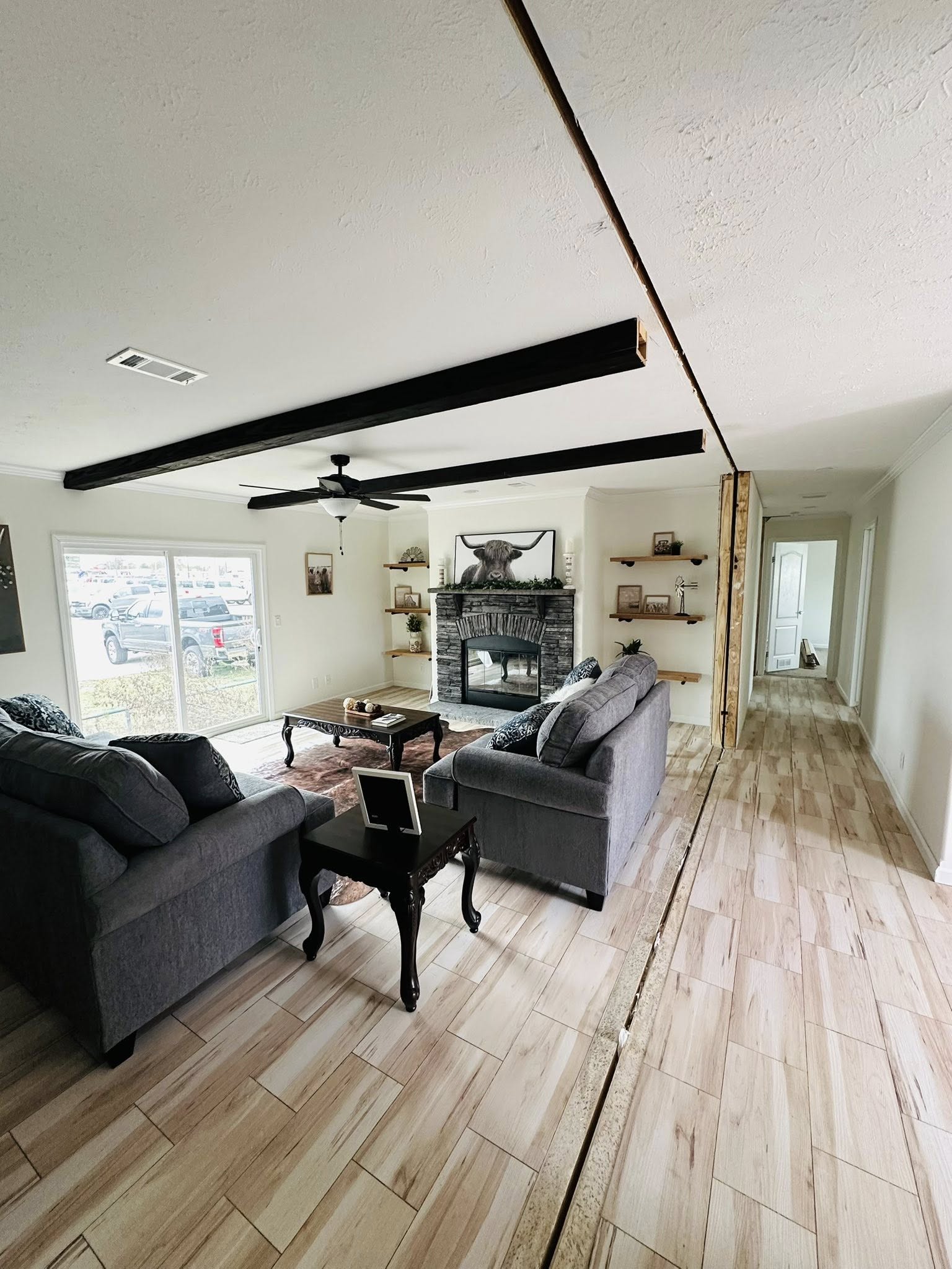 Spacious living room with gray sofas, a wooden coffee table, and a brick fireplace. Shelves display decor. Light wood flooring and large windows. Cozy and inviting.