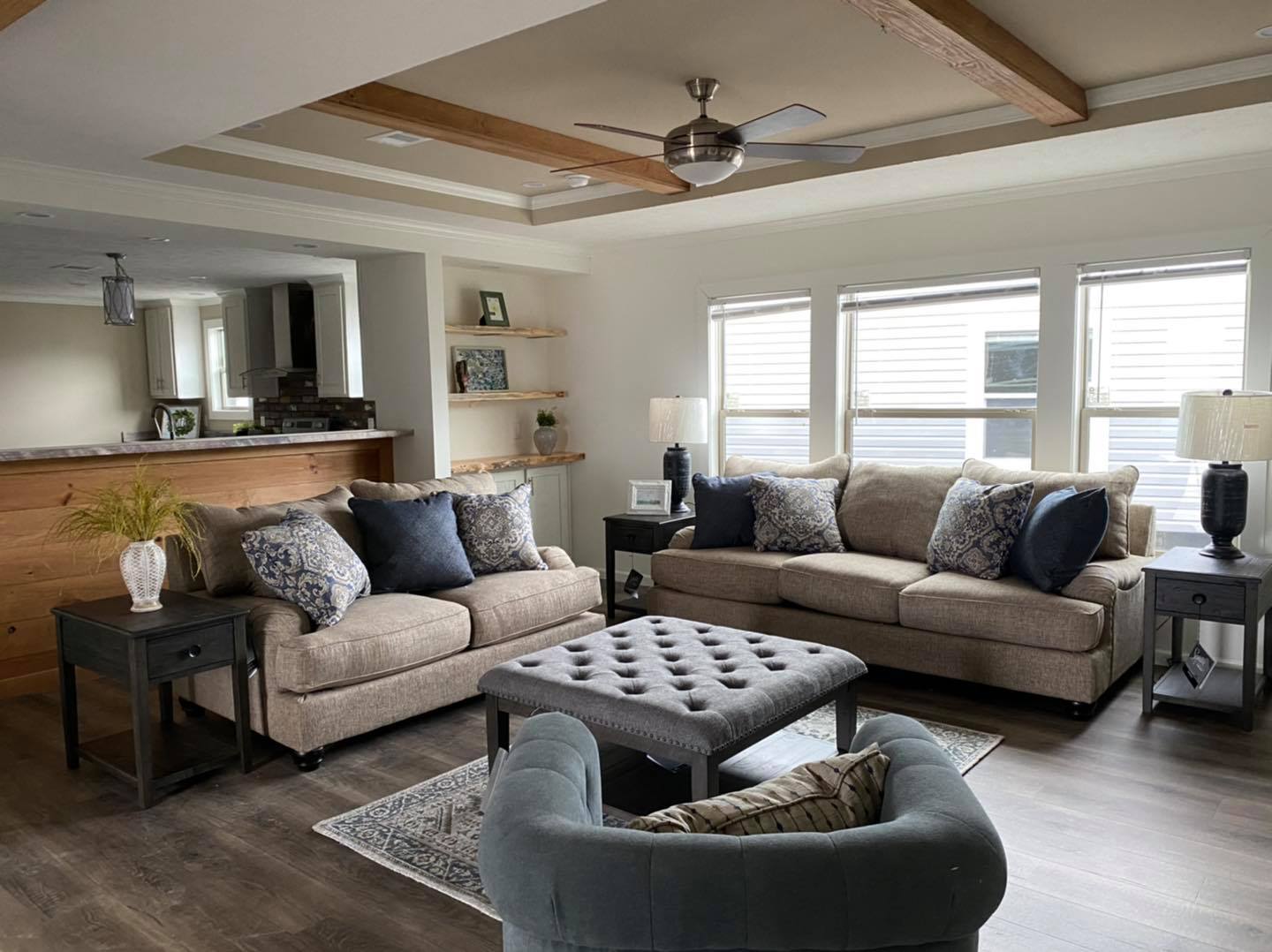 Spacious, modern living room with neutral tones. Two beige sofas and a tufted ottoman, four windows, ceiling fan, and dark wood side tables create a cozy atmosphere.