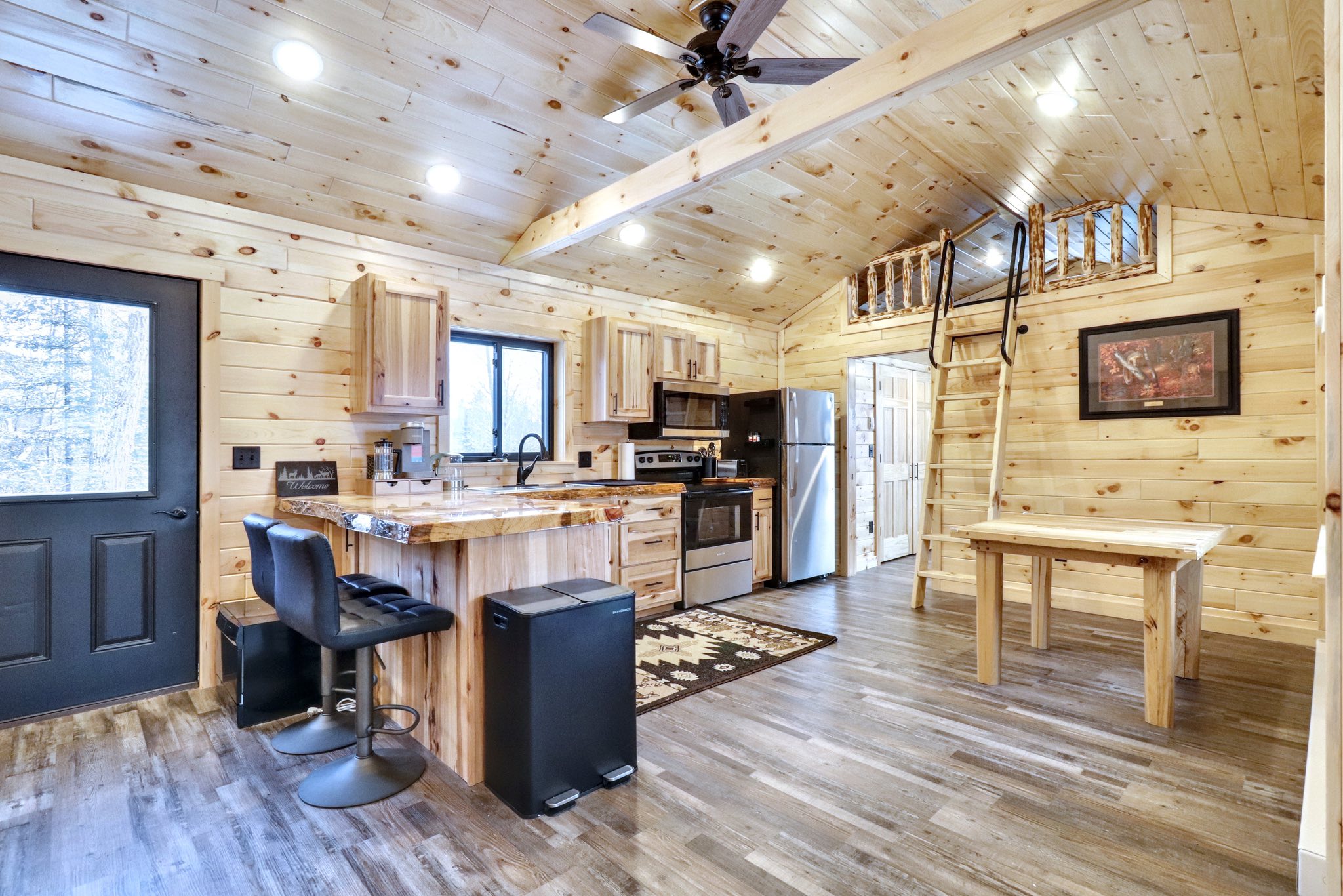Cozy log cabin interior with wood-paneled walls and ceiling, modern kitchen, island with stools, loft with ladder, and rustic decor. Warm, inviting atmosphere.