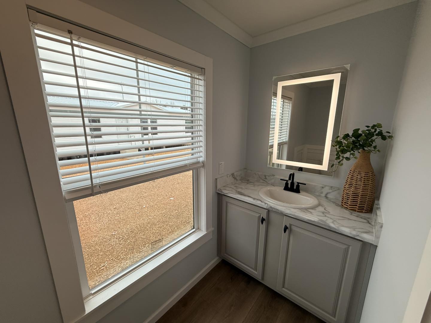 A small modern bathroom with a window on the left, beige blinds, and a wooden floor. A marble sink counter with a lit mirror and plant on the right.