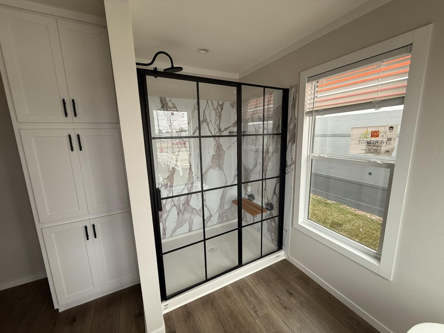 Modern bathroom with a glass shower enclosure featuring marble-patterned walls and black grid lines, beside white storage cabinets and a large window. Warm tone.