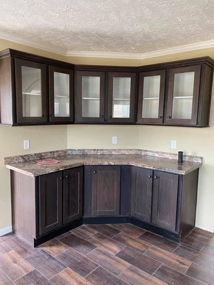Corner kitchen cabinets with dark wood finish and glass upper doors. Brown granite countertop, beige walls, and wood-patterned floor create a warm, cozy feel.