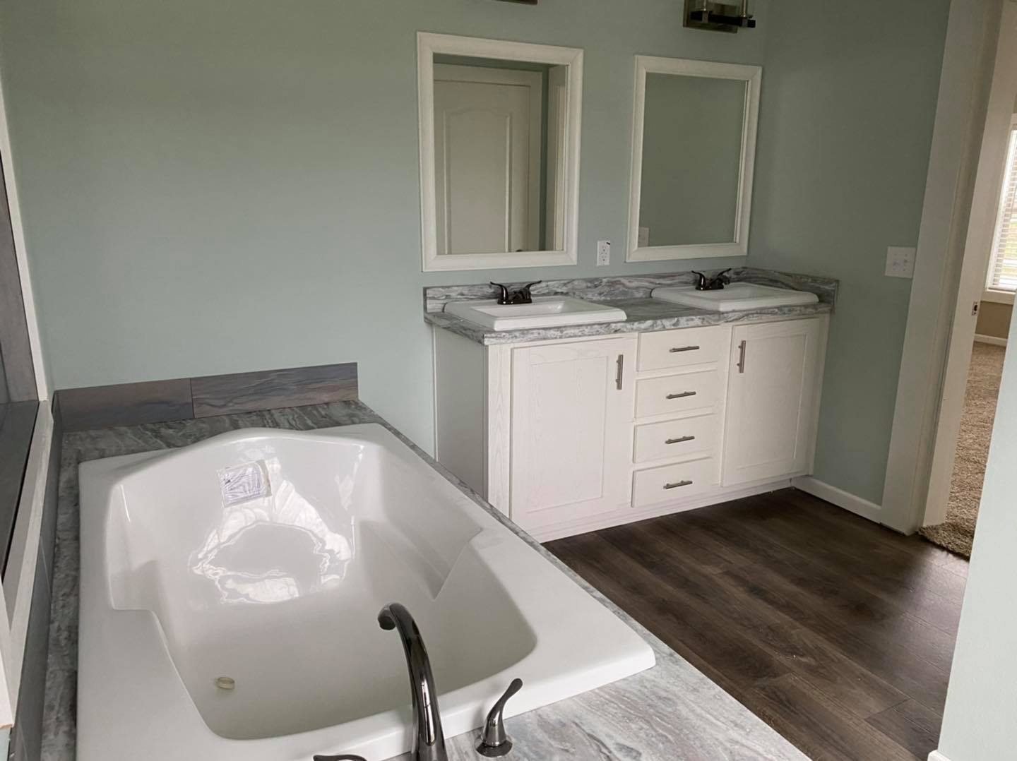 Modern bathroom with a marble-countered double sink and mirrors. A white bathtub with a sleek, black faucet is adjacent on dark wood flooring. Calm ambiance.