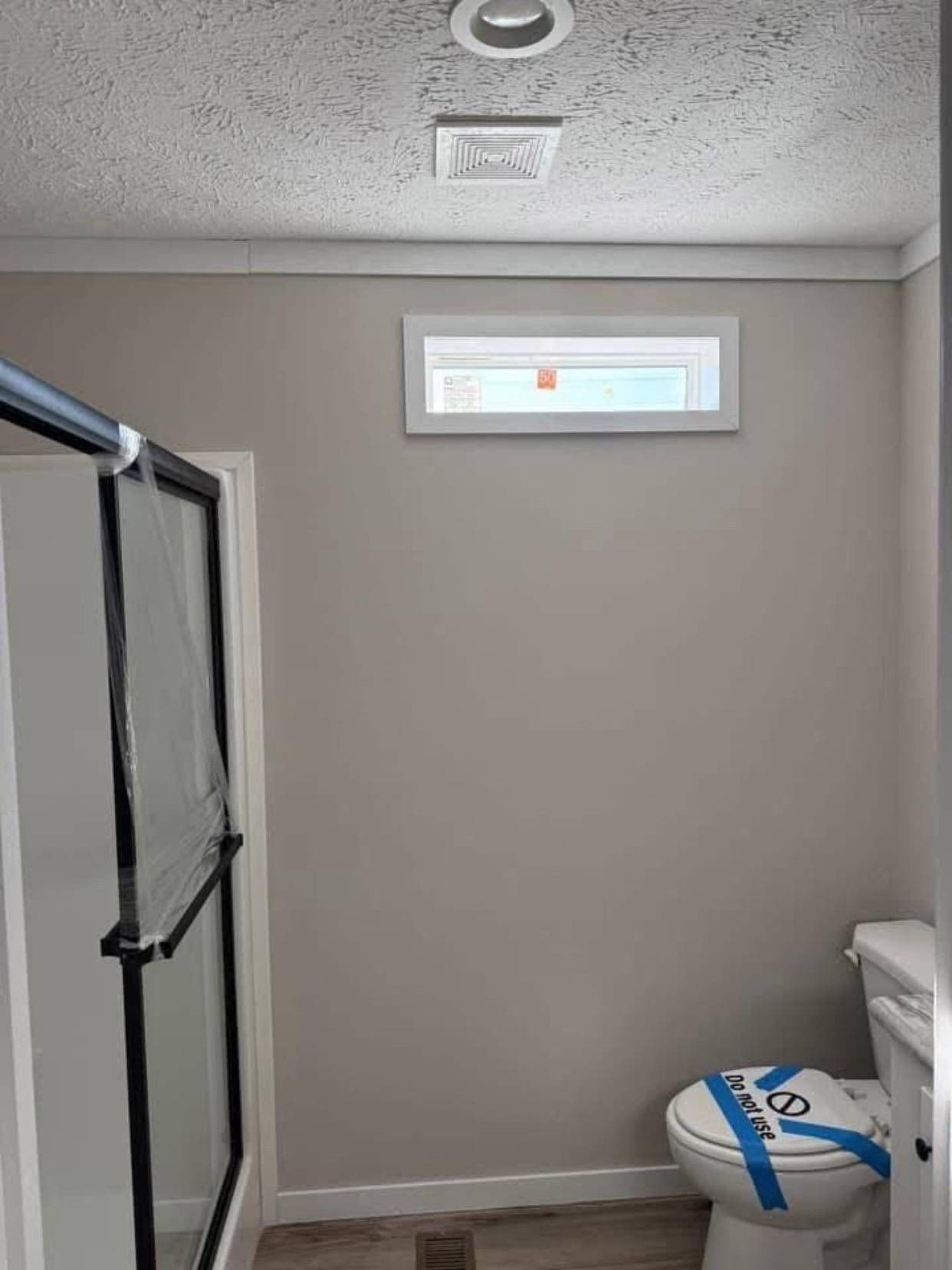 Small bathroom with gray walls, a frosted glass shower door on the left, and a toilet on the right marked with "Do not use" tape. Natural light enters through a high, narrow window.