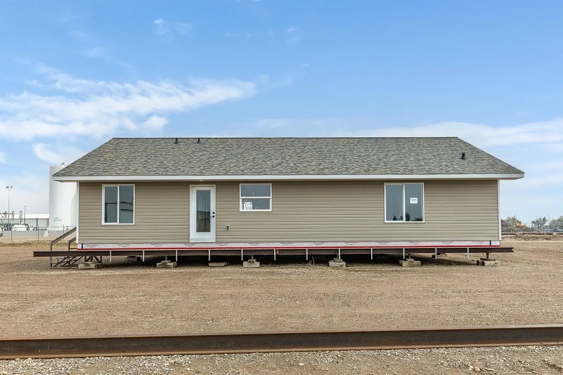 Single-story prefab house with beige siding and dark roof, elevated on a dirt lot. A clear blue sky in the background creates a serene atmosphere.