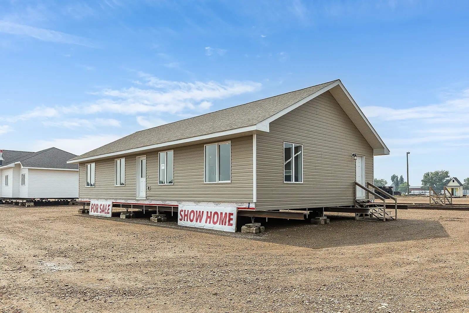 Single-story, beige modular home on an empty dirt lot under a clear blue sky. Signs read "For Sale" and "Show Home," with stairs leading to the entrance.