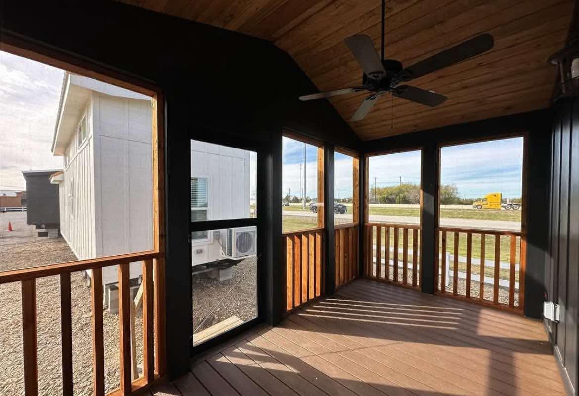 Wooden porch with ceiling fan, featuring large screened windows. Sunlight casts shadows on the floor. A white building and truck are visible outside.