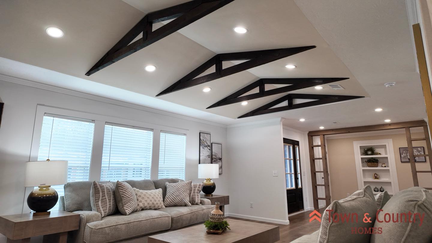 Modern living room with a cozy beige sofa, geometric black ceiling beams, soft lighting, and decorative accents. Bright and inviting ambiance.