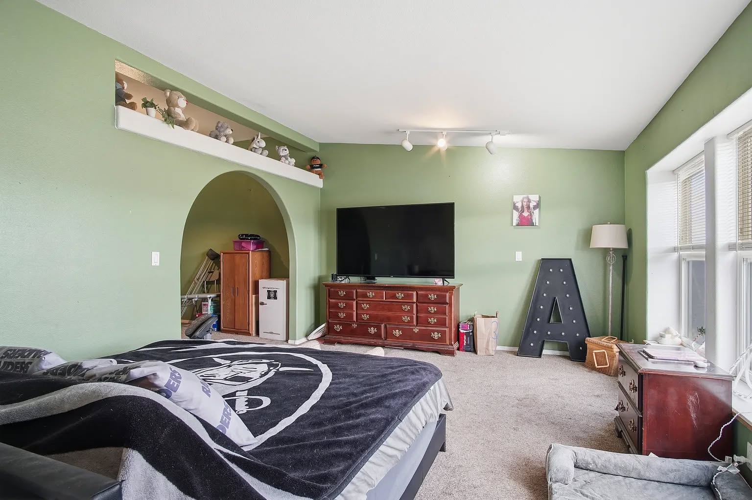 Spacious bedroom with green walls, featuring a large TV on a dresser, a bed with a sports-themed blanket, and a decorative letter "A" on the floor.