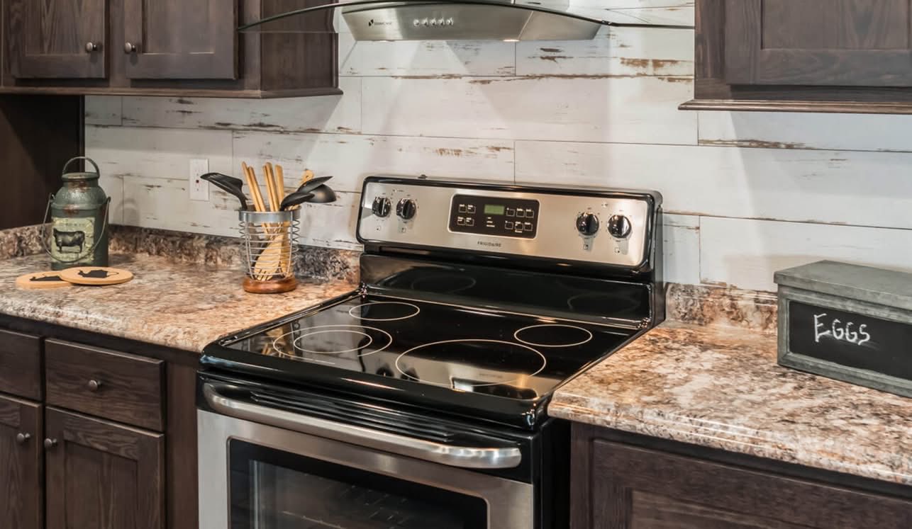 A modern kitchen with a stainless steel electric stove, dark wood cabinets, marbled countertops, and rustic accents like a tin jar and wooden utensils.