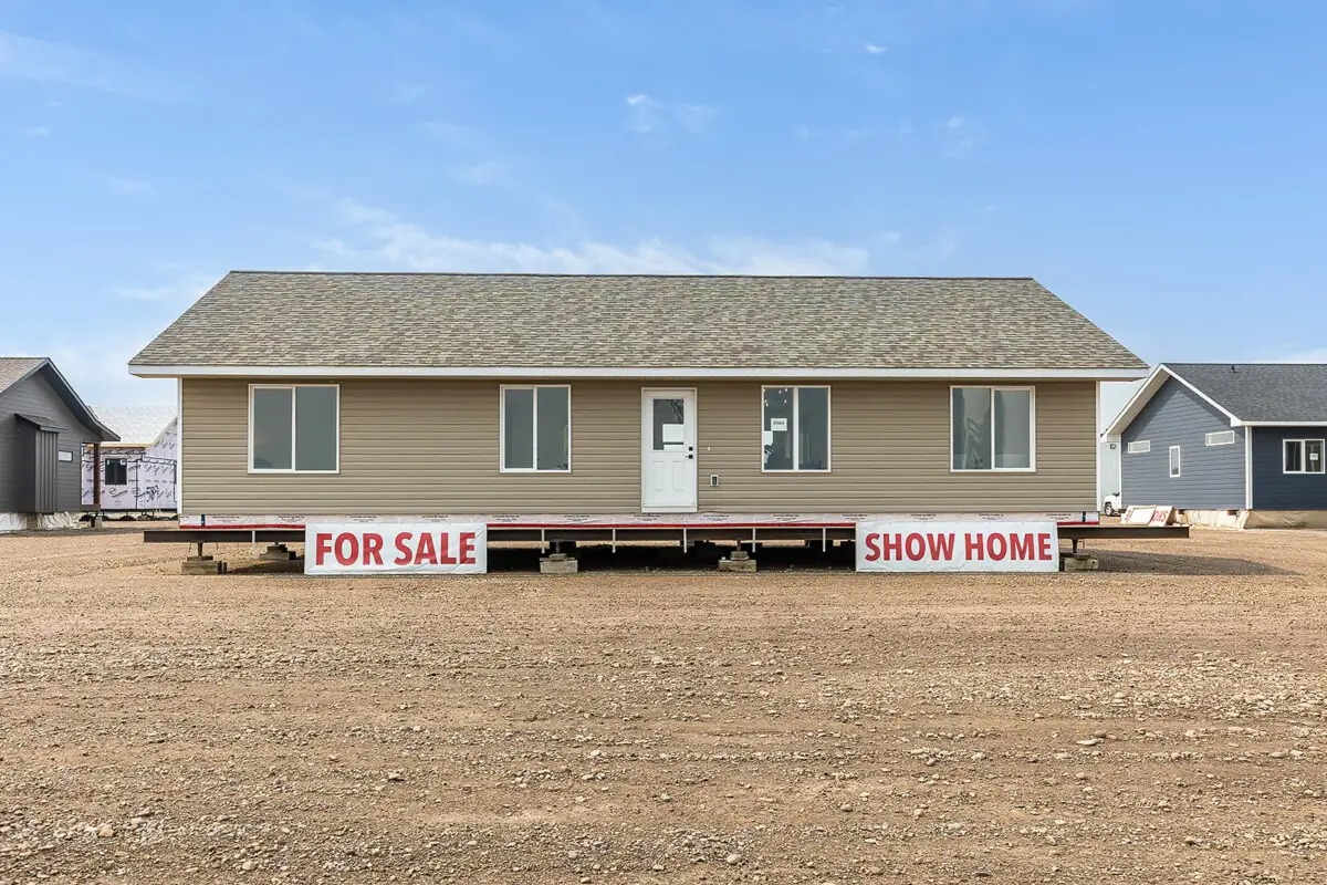 Single-story tan house labeled "For Sale" and "Show Home," on gravel lot under blue sky, conveying availability and invitation to view.