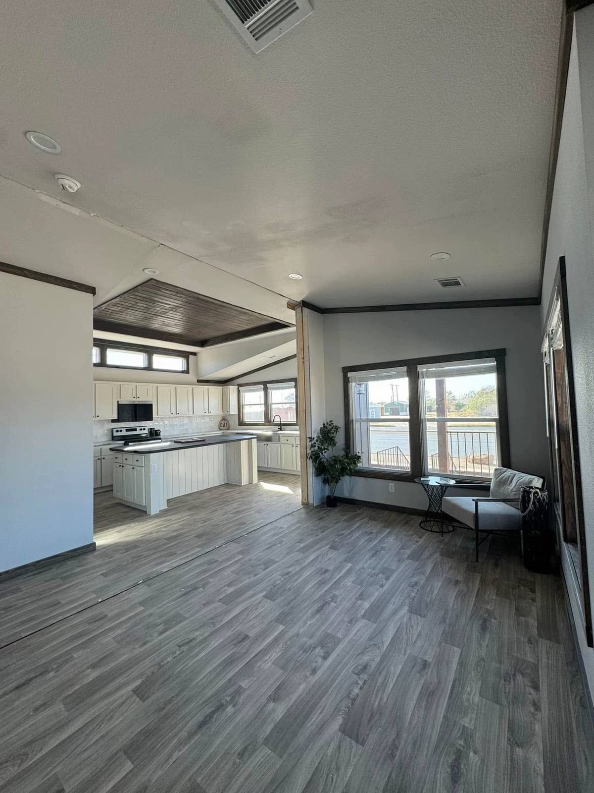 Open living space featuring light wood flooring, a modern kitchen with white cabinets, and large windows. A chair with a small table and plant convey a cozy feel.