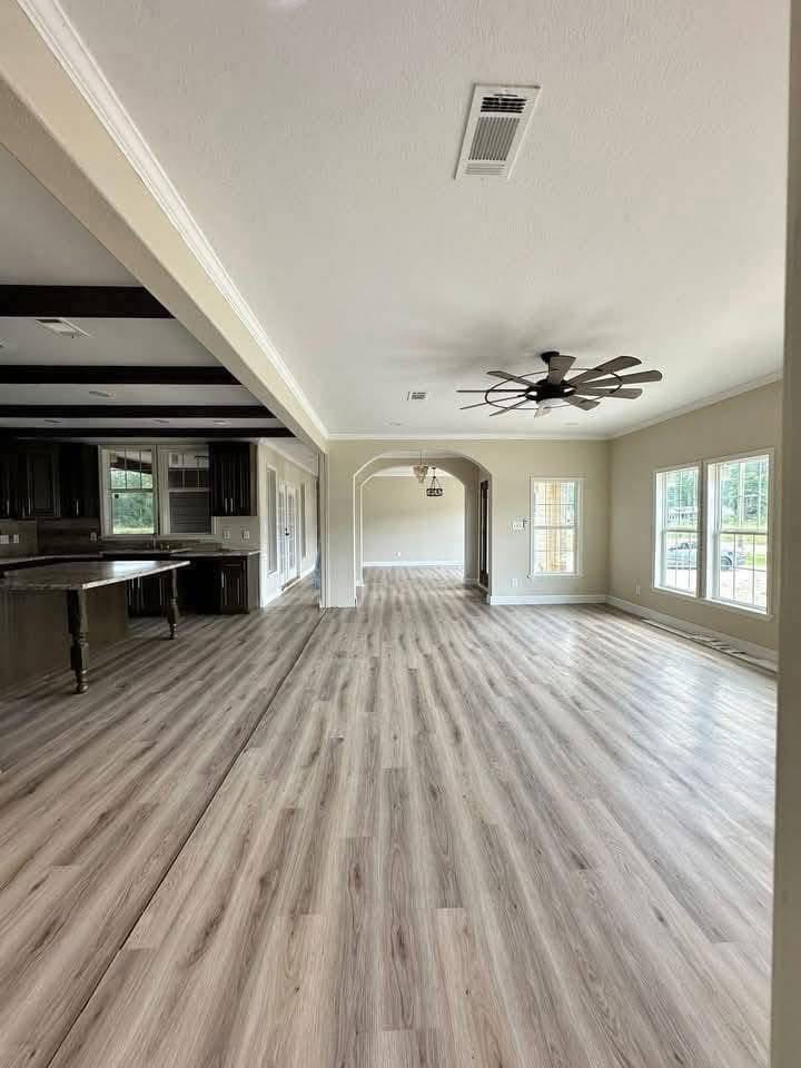 Spacious open-plan room with light wood flooring, large windows, modern ceiling fan, and visible kitchen area. The atmosphere feels bright and airy.