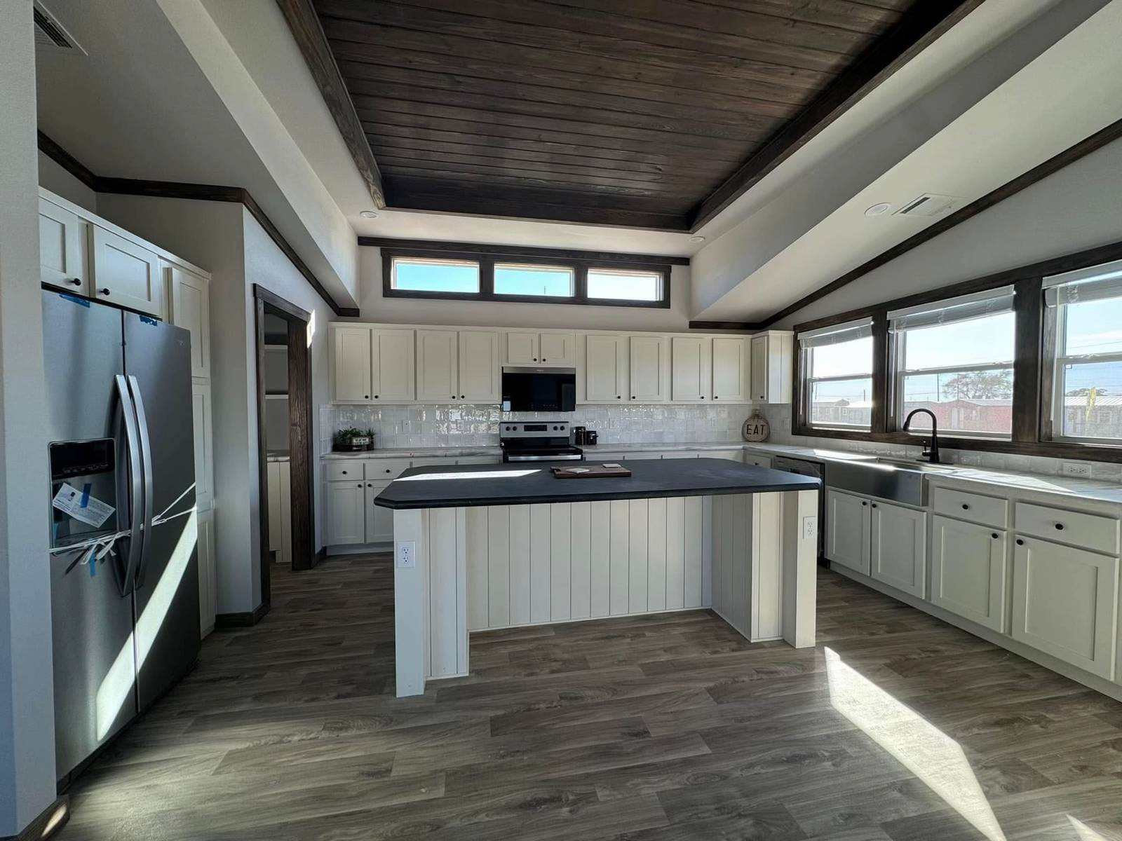 Modern kitchen with dark wood ceiling, white cabinets, and a large island featuring a gray countertop. Stainless steel fridge and sunlight streaming in.