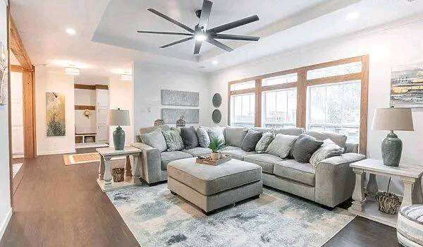 Spacious living room with a gray sectional sofa, large ottoman, and stylish decor. Natural light from multiple windows enhances a cozy ambiance.