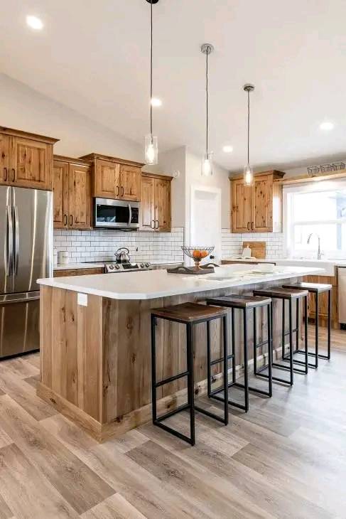 Spacious kitchen with wooden cabinets, an island with four black stools, stainless steel appliances, and a bright, airy feel. Hanging pendant lights add warmth.