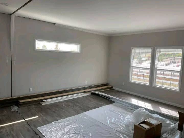 A room with light gray walls and large windows. Wooden floorboards and paint sheets are laid out, suggesting ongoing renovations. Bright, airy atmosphere.
