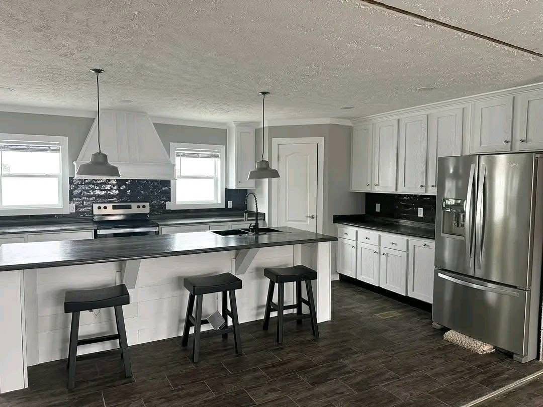 Modern kitchen with gray tones, featuring a large island with three stools, pendant lights, stainless steel appliances, and white cabinetry. Bright and sleek.
