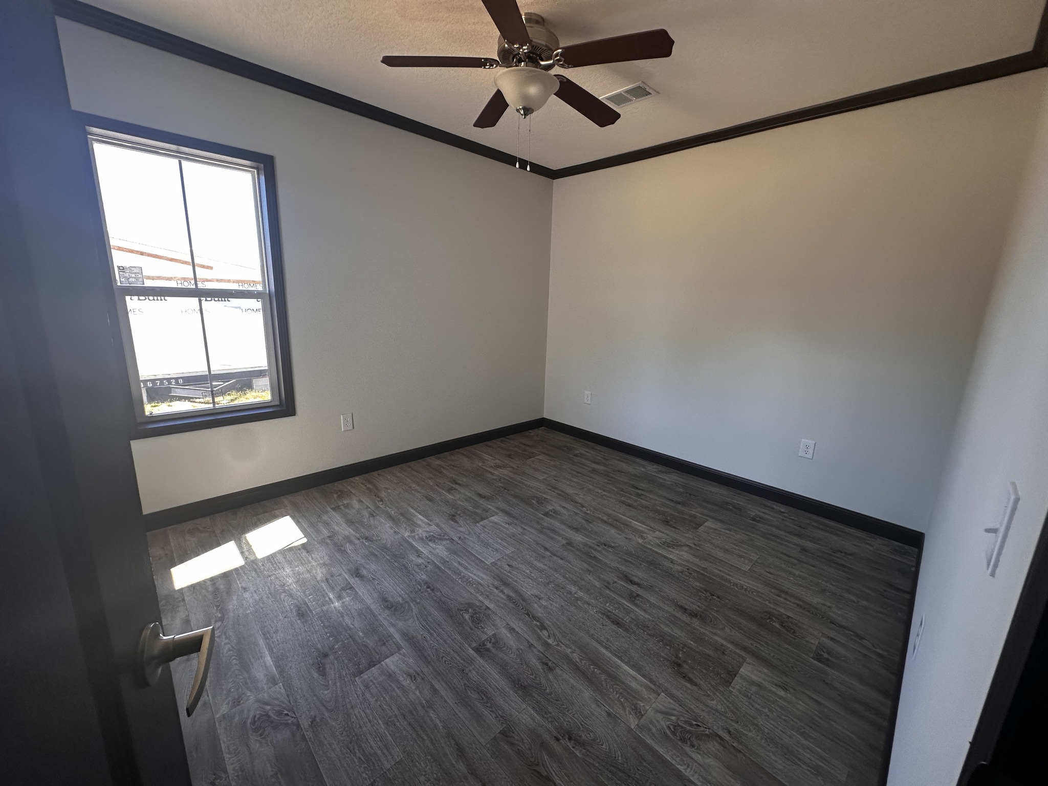 Empty room with gray wood flooring, white walls, and dark trim. A ceiling fan is mounted above, and light streams through a window on the left.