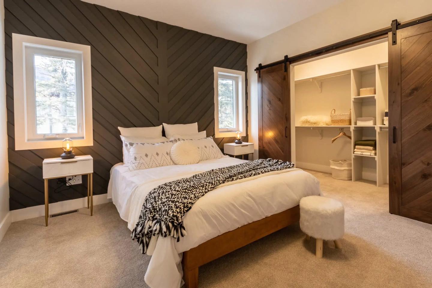 Cozy bedroom with white bedding, plush pillows, and a patterned throw. Features a dark accent wall, two windows, soft lighting, and open wooden closet doors.
