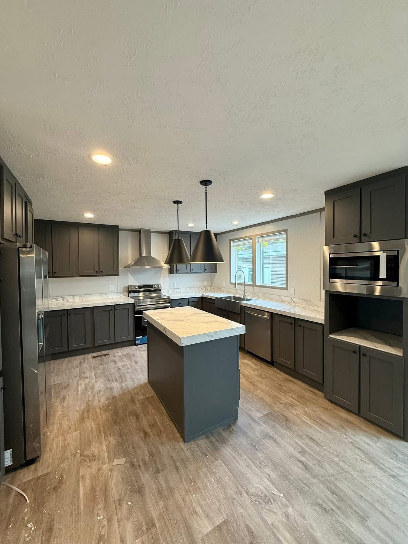 Modern kitchen with dark gray cabinets, marble countertops, island, and stainless steel appliances. Pendant lights and wood flooring add warmth.