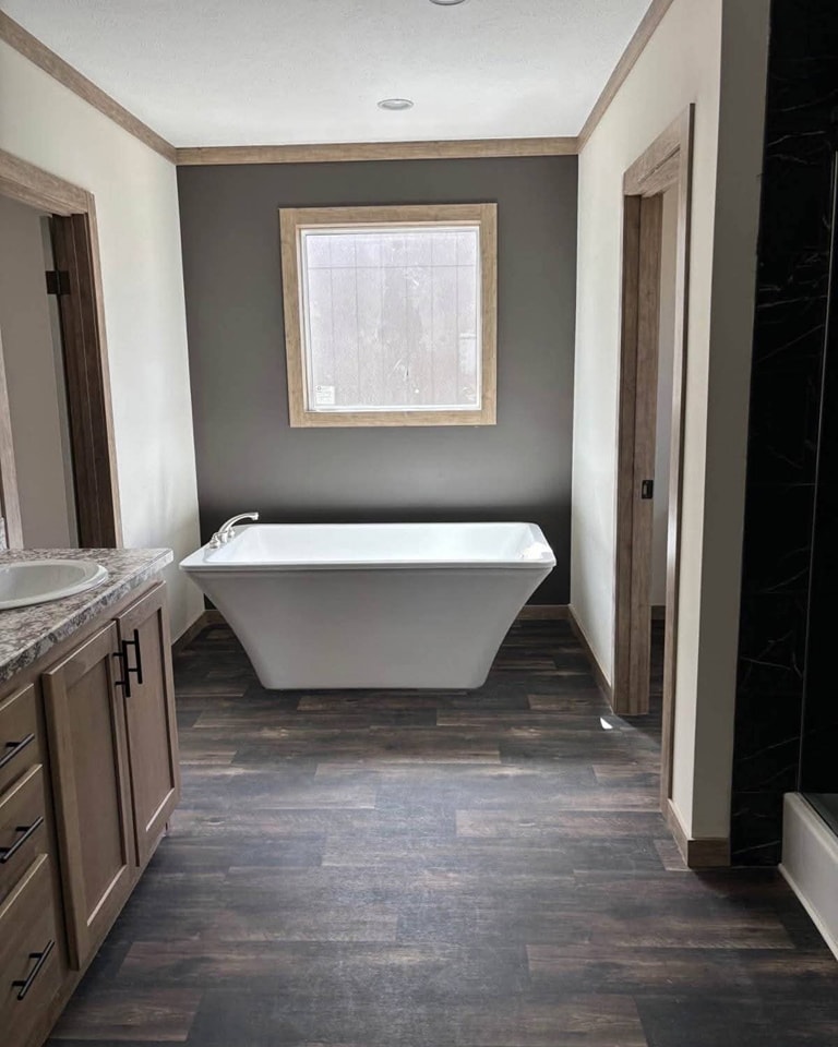Minimalist bathroom with a central white freestanding bathtub, dark wood floors, and a gray accent wall with a large window, creating a serene atmosphere.