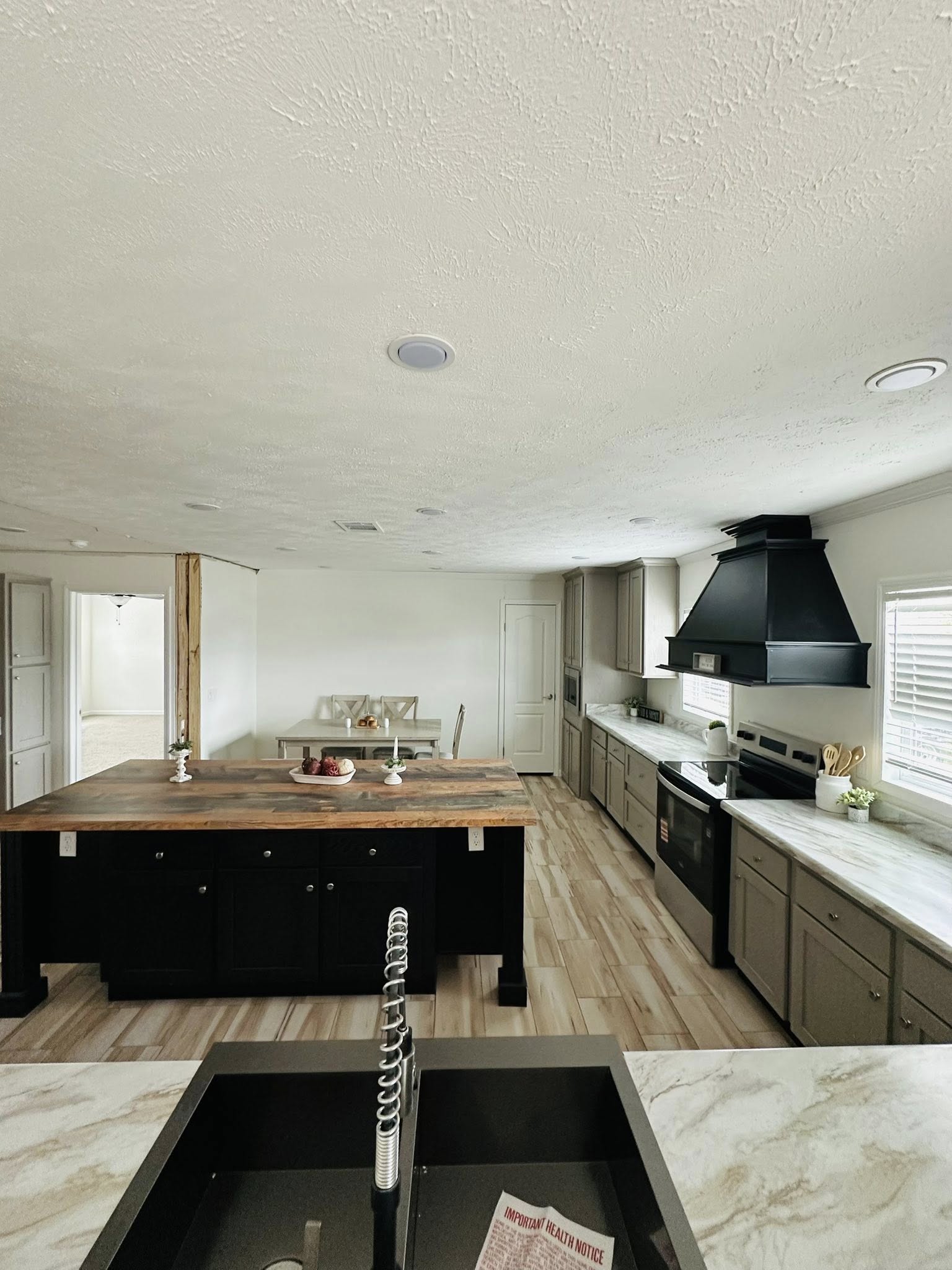 Spacious kitchen with white ceiling and wooden floor. Features a large wooden island, modern black appliances, gray cabinets, and a small dining area. Cozy and inviting.
