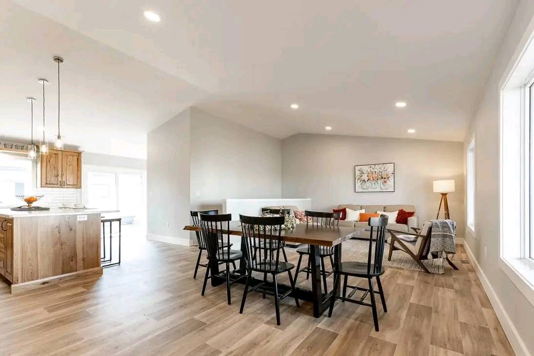 Spacious open-concept living area with wooden floors, featuring a dining table and black chairs. In the background, a cozy lounge with a sofa, colorful cushions, and a standing lamp creates a warm, inviting atmosphere.