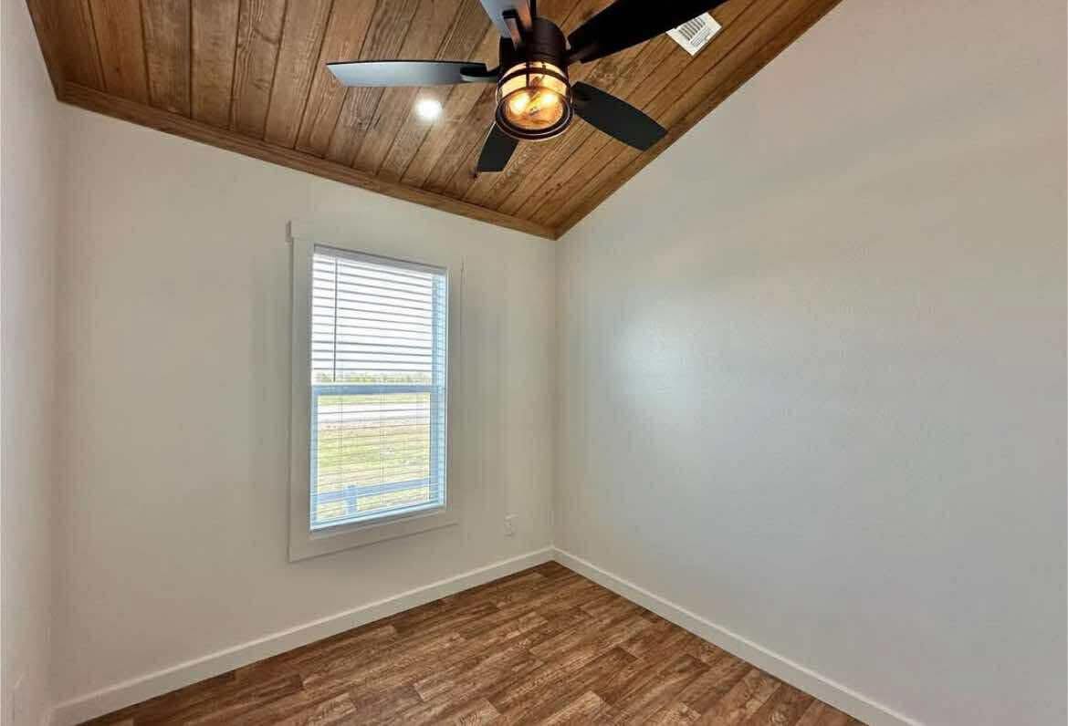 Small room with white walls, wooden ceiling, and flooring. A modern black ceiling fan hangs overhead. Light from a window softly illuminates the space.