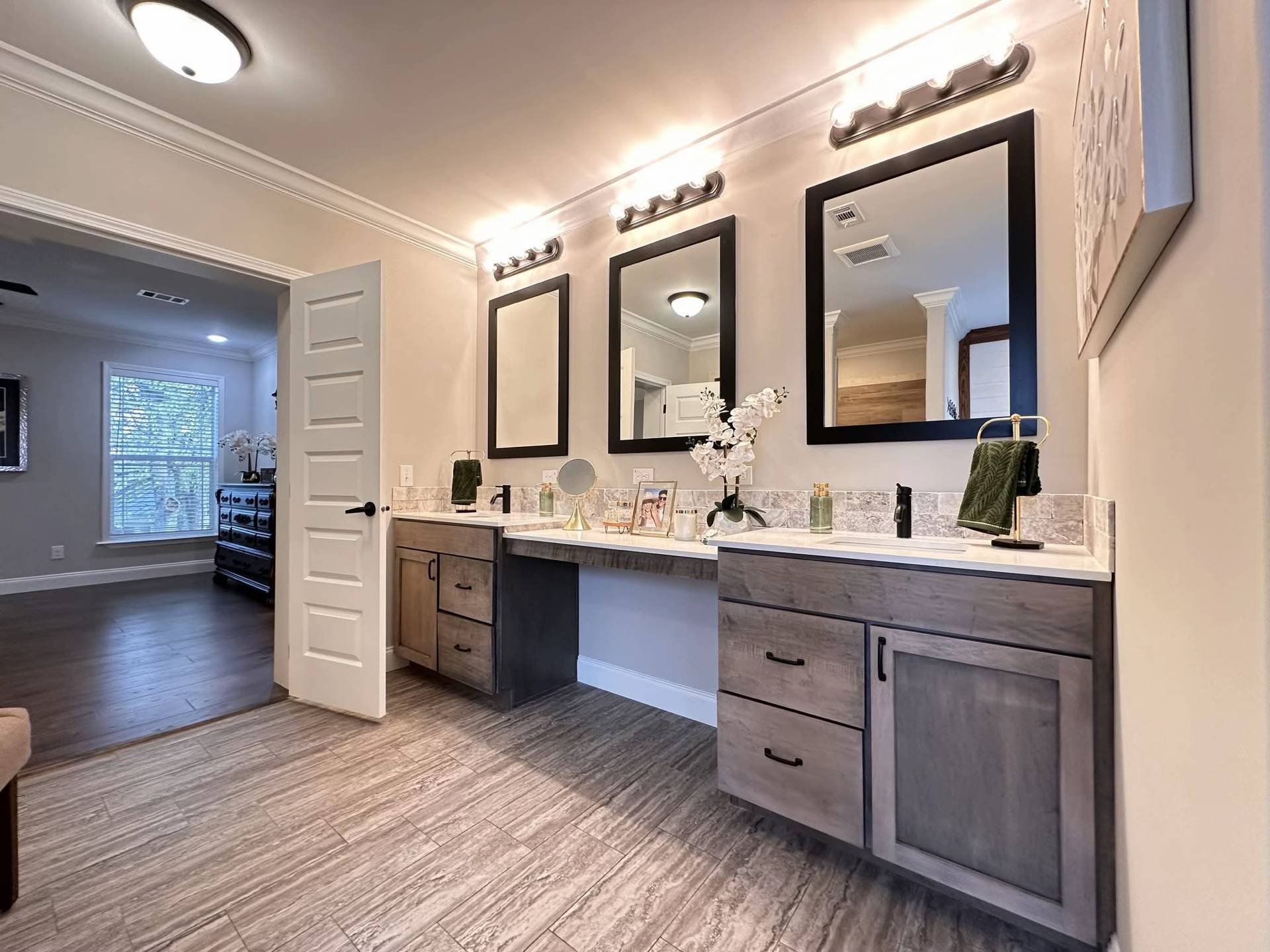 Modern bathroom with dual wooden vanities, three large mirrors, and soft lighting. Decorative orchids add elegance, while an open door leads to a cozy room.