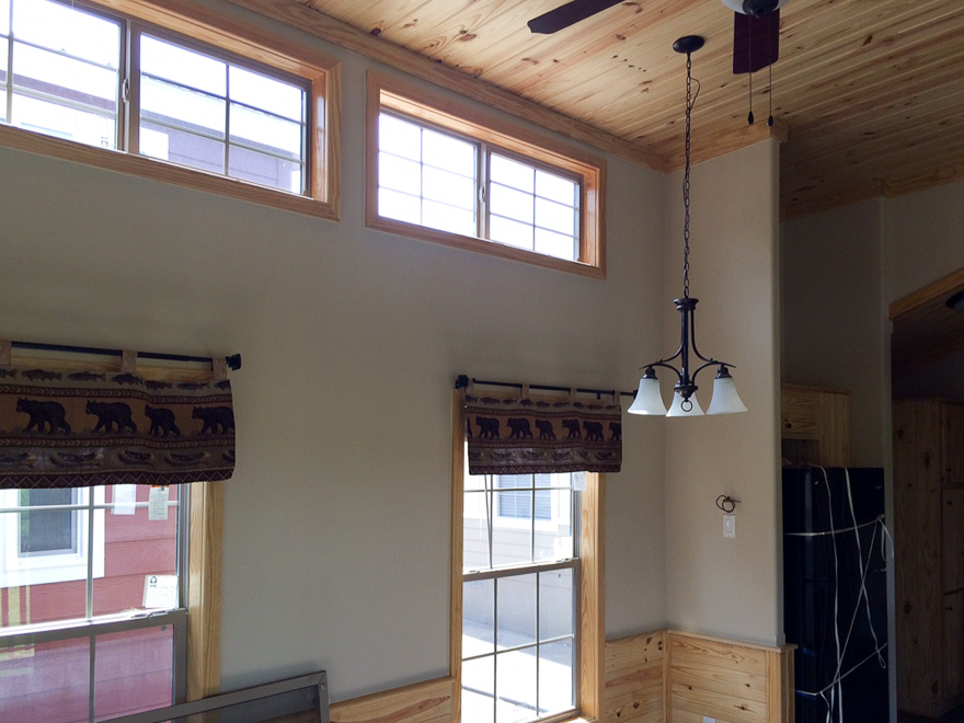 Cozy room with light wood ceiling and walls. Large windows have bear-patterned curtains. A black chandelier hangs from the ceiling, adding rustic charm.