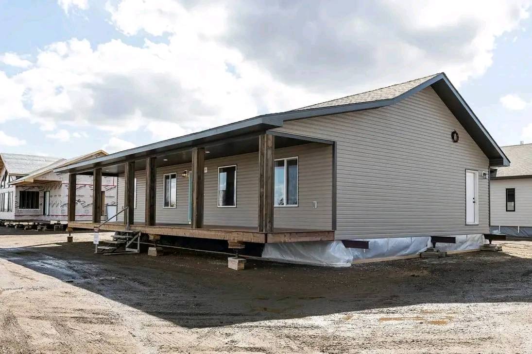 A modular home with gray siding and a front porch sits on a dirt lot under a partly cloudy sky. It is elevated on blocks, suggesting ongoing construction.