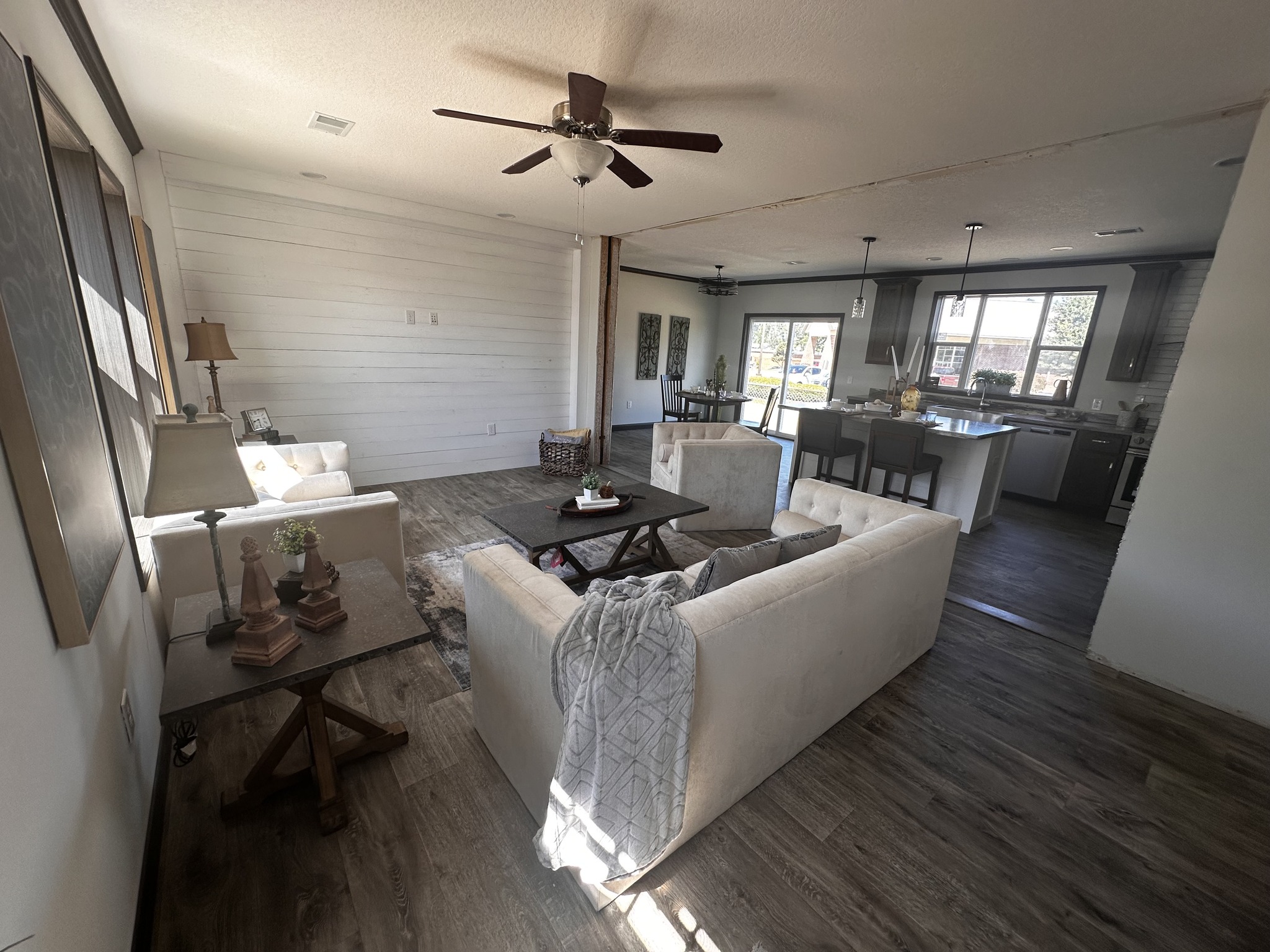 Spacious living room with beige sofas, wooden coffee table, and ceiling fan, leading to an open kitchen and dining area. Bright, welcoming ambiance.