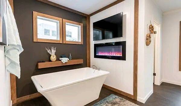 Modern bathroom with a sleek white tub, dual windows, and a wall-mounted electric fireplace. Wooden accents add warmth and a touch of elegance.