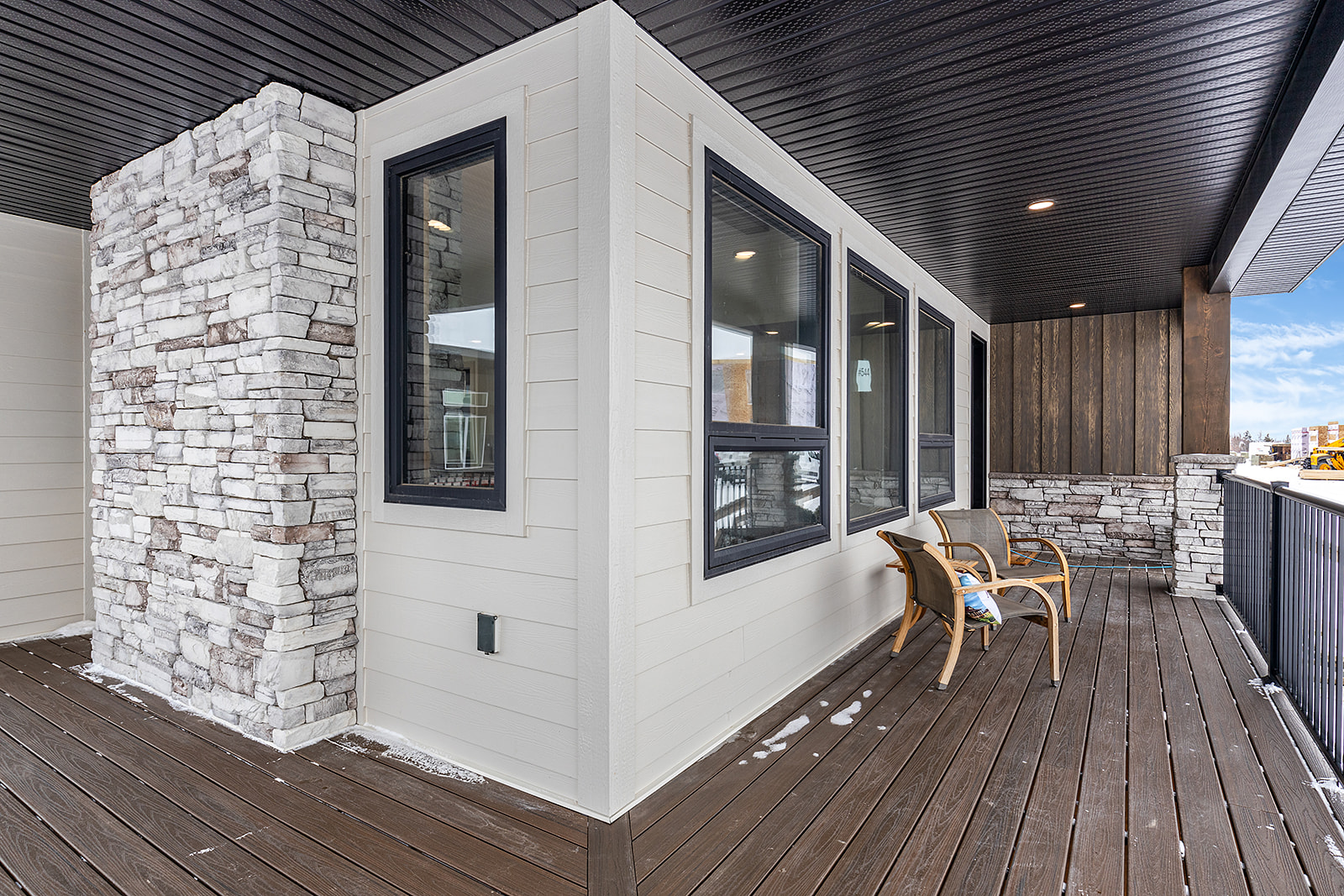 Modern balcony with wooden flooring and stone walls, featuring black-framed windows. Two wooden chairs offer a cozy outdoor seating area.