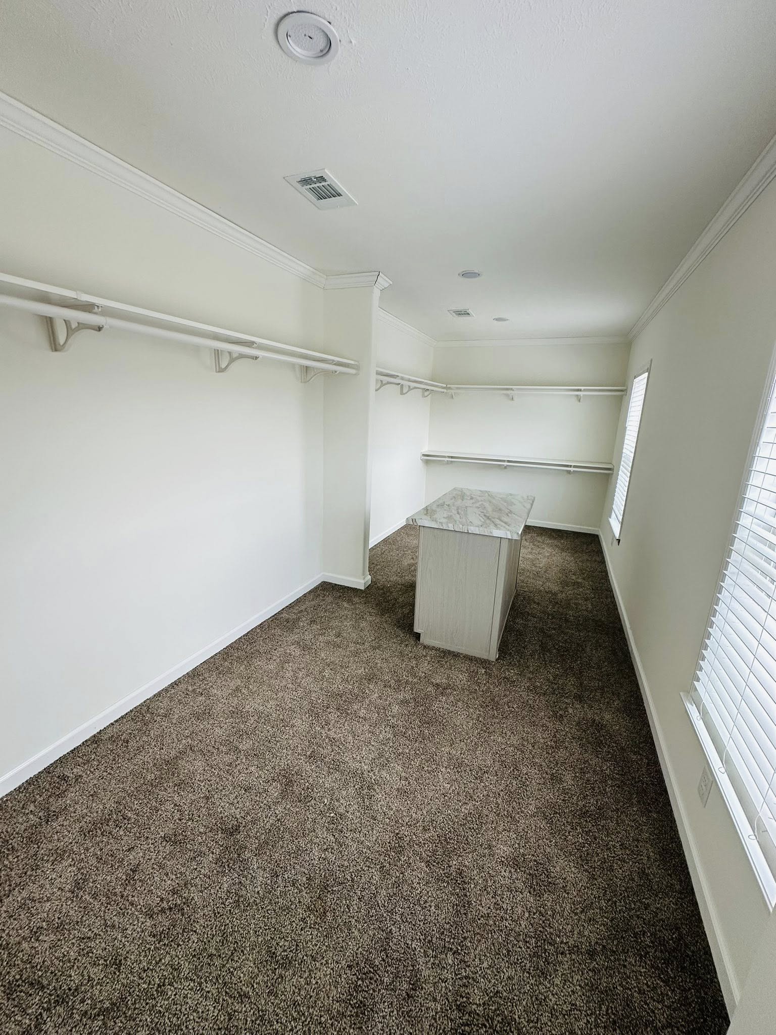 Spacious walk-in closet with beige carpet, white walls, and ceiling. It features long shelves on each side and a central island with a marble countertop. Natural light from a window creates a serene, organized feel.