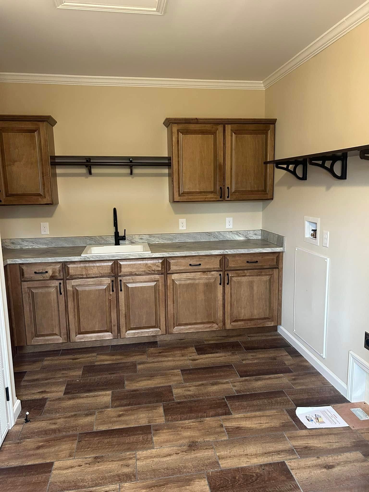 A small laundry room with wood cabinets and tiled floor. The room has beige walls, a countertop with a sink, and black faucet. Shelves are on the walls.